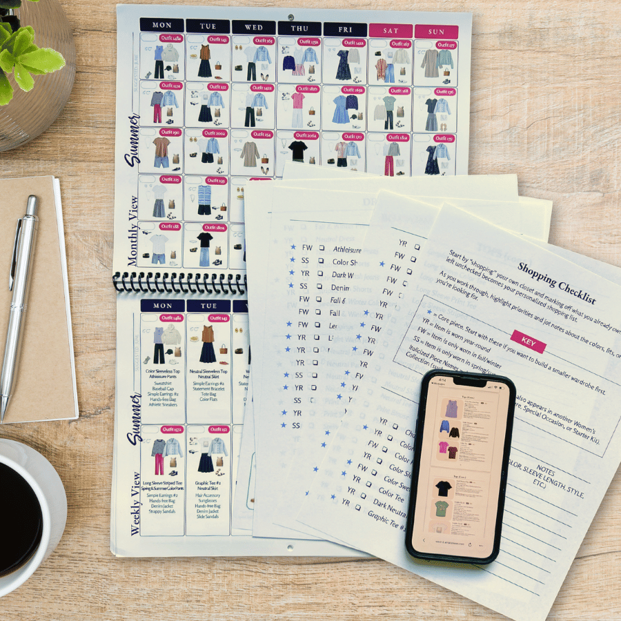 Spiral-bound Frump Fighters /what-to-Wear outfit calendar open to a summer monthly and weekly view, showing daily outfit combinations with clothing images and outfit numbers. Printed shopping checklist pages with season codes and checkboxes fan out beside it, next to a phone displaying the digital calendar, a coffee cup, notebook, and pen on a wood desk.